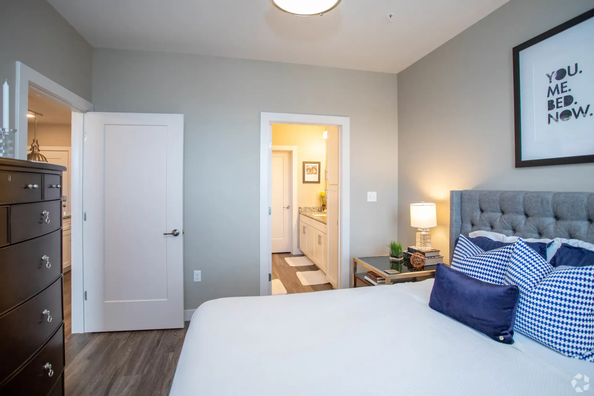 Bedroom with bed, dresser, and bathroom door; neutral color palette with blue accents at Chroma, offers apartments near The Grove in St Louis.