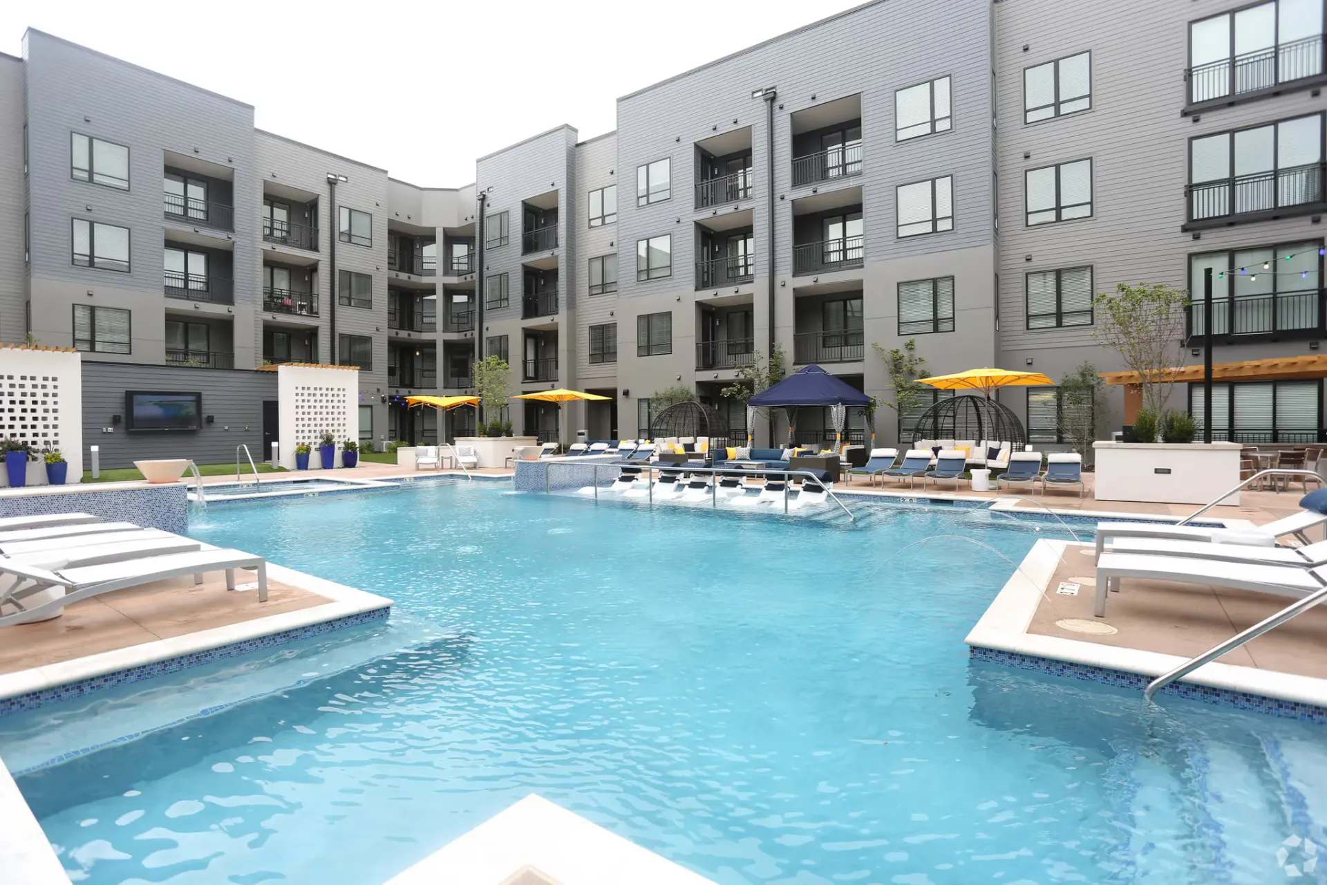 The swimming pool in front of the modern apartment complex at Chroma offers STL apartments for rent, featuring multiple stories, loungers, and blue umbrellas.
