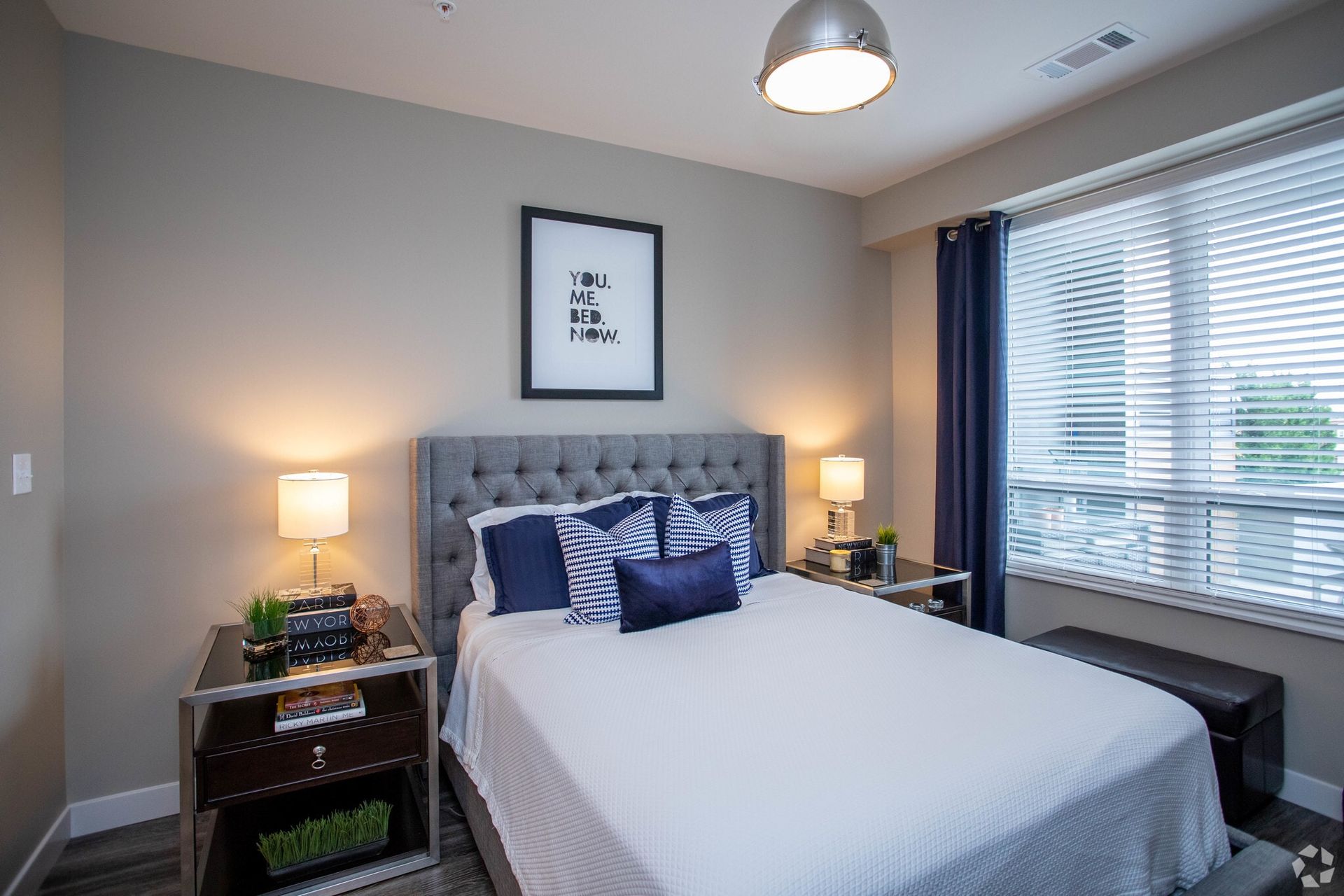 Bedroom with a gray upholstered bed, nightstands, and window with blinds at Chroma, offers apartments in The Grove.