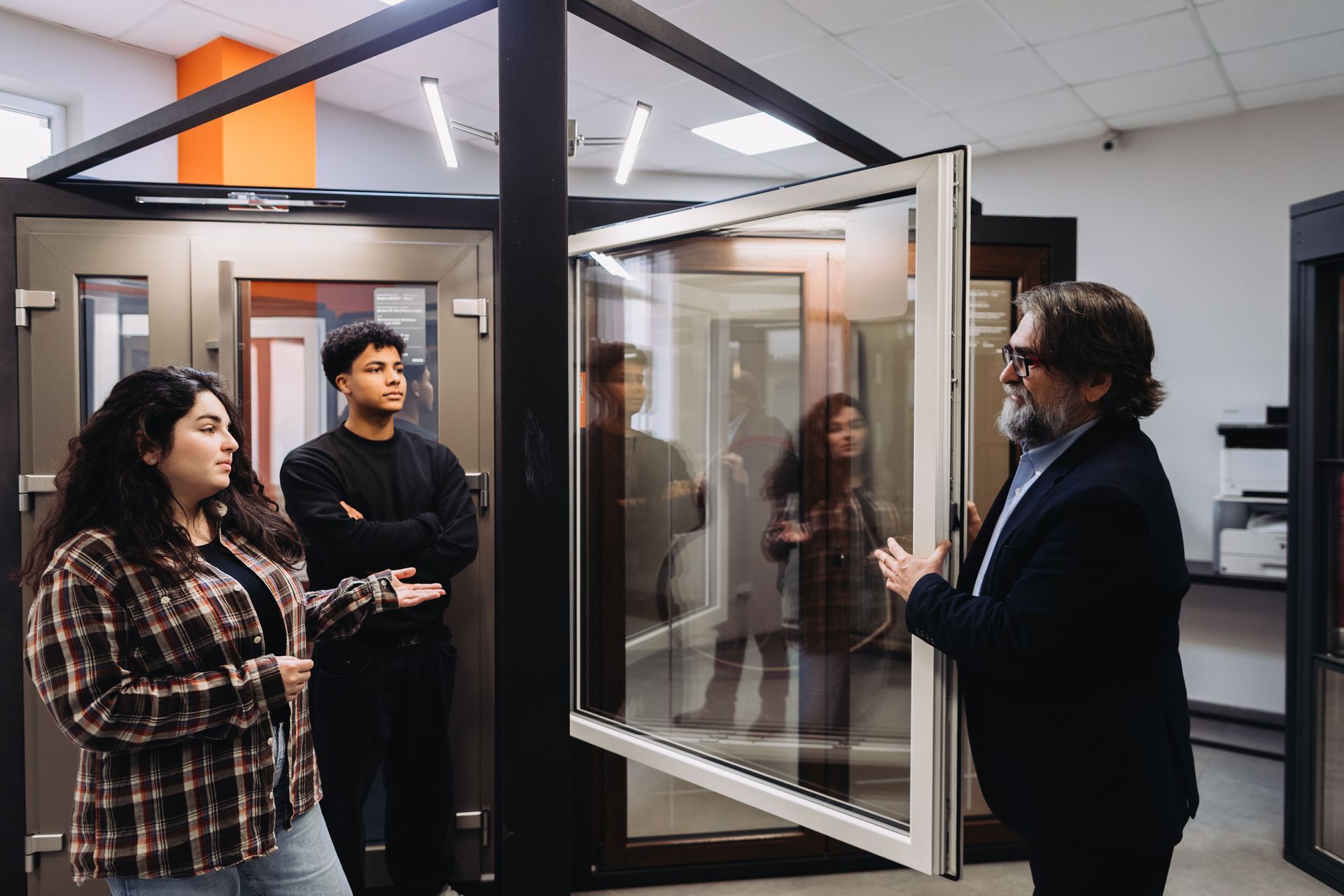 Three people listen as a man demonstrates a glass office booth in a modern workspace. Three people listen as a man demonstrates a glass office booth in a modern workspace.