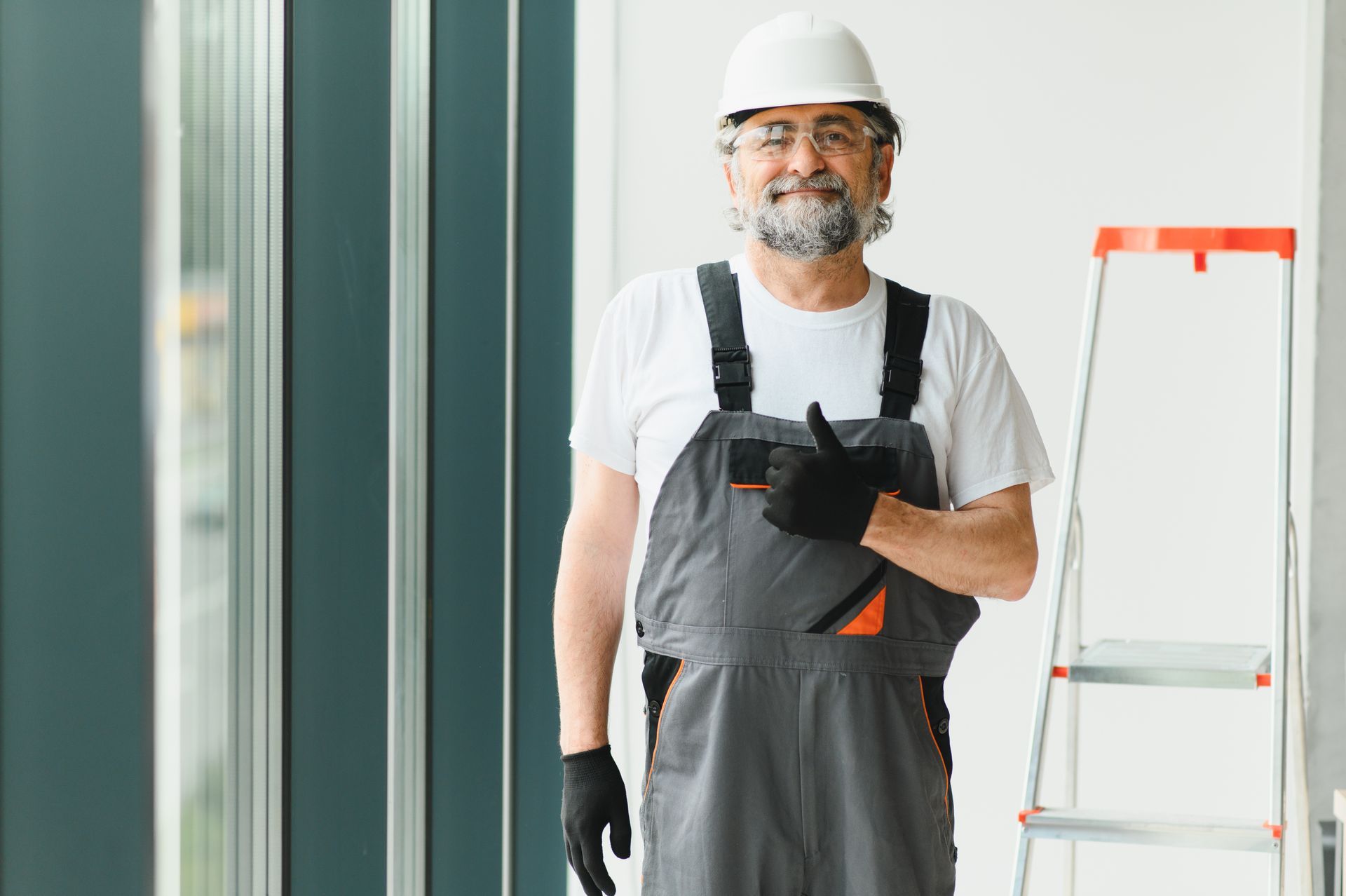 Construction worker in hard hat and overalls standing beside a ladder indoors Construction worker in hard hat and overalls standing beside a ladder indoors