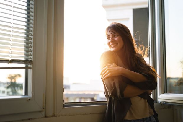 Woman standing by a window, smiling in warm sunlight with blinds and city view behind her Woman standing by a window, smiling in warm sunlight with blinds and city view behind her