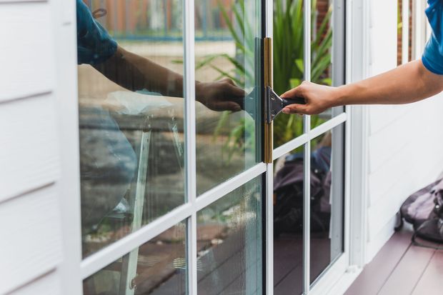 Hand reaching through glass patio door to open black handle, with white siding and deck railing nearby Hand reaching through glass patio door to open black handle, with white siding and deck railing nearby