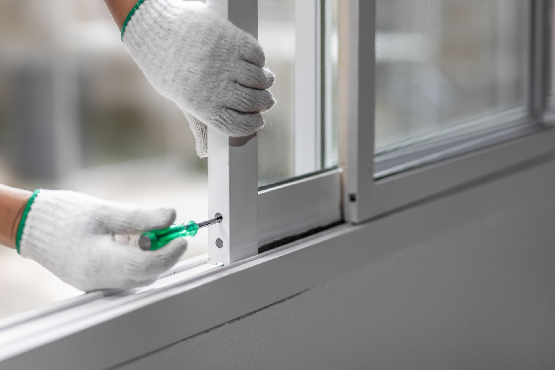 Gloved hands use a screwdriver to install a white window frame latch. Gloved hands use a screwdriver to install a white window frame latch.