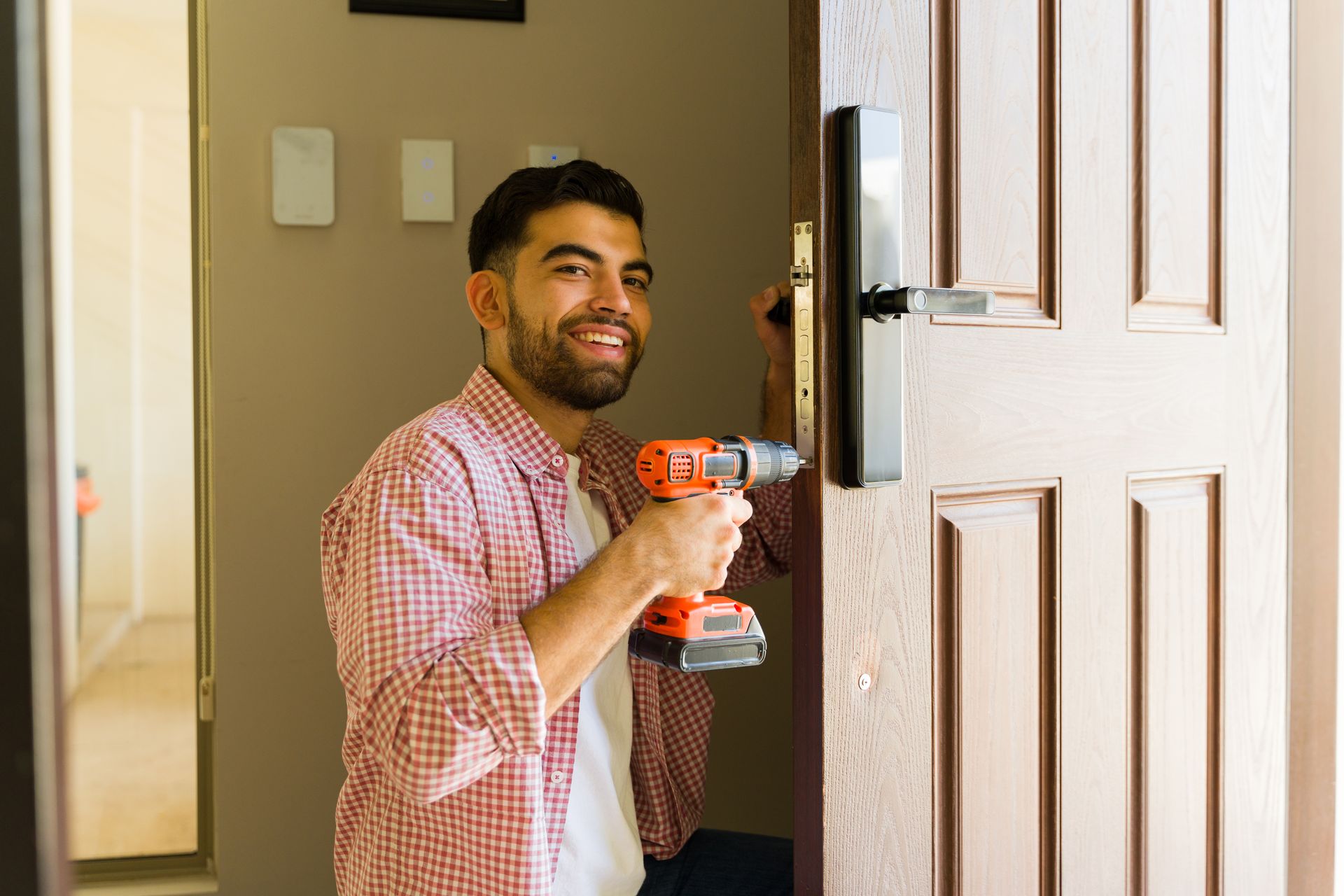 Man holding an orange power drill while opening a white front door inside a home Man holding an orange power drill while opening a white front door inside a home