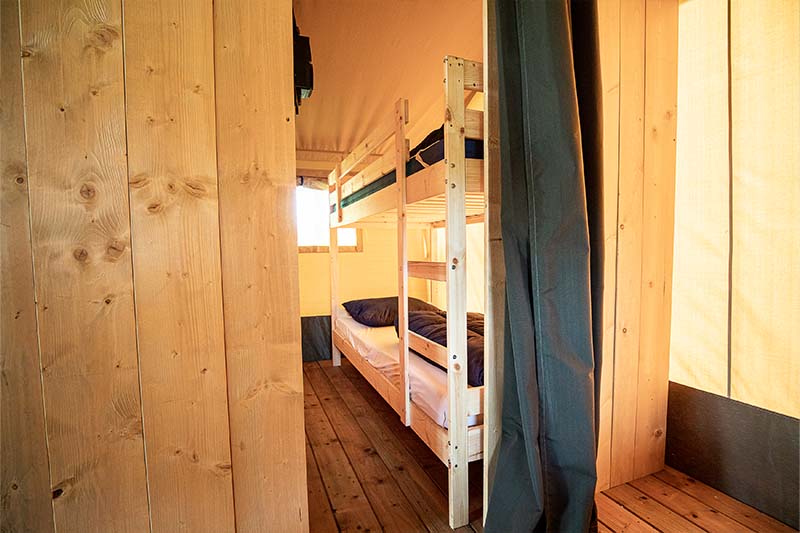 Houten hut met stapelbedden, een smalle gang, een stapelbed en warm zonlicht dat door een klein raam naar binnen schijnt.