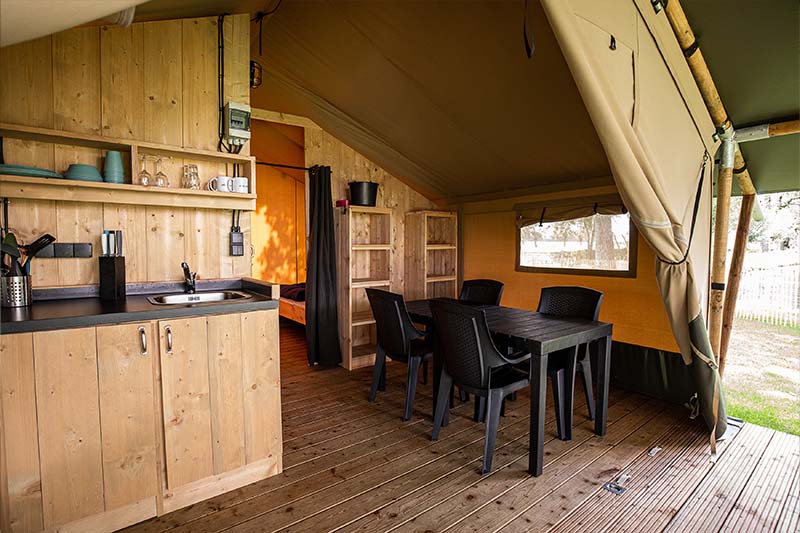 Interieur van een safaritent met houten keuken, eettafel en zwarte stoelen op een houten terras.