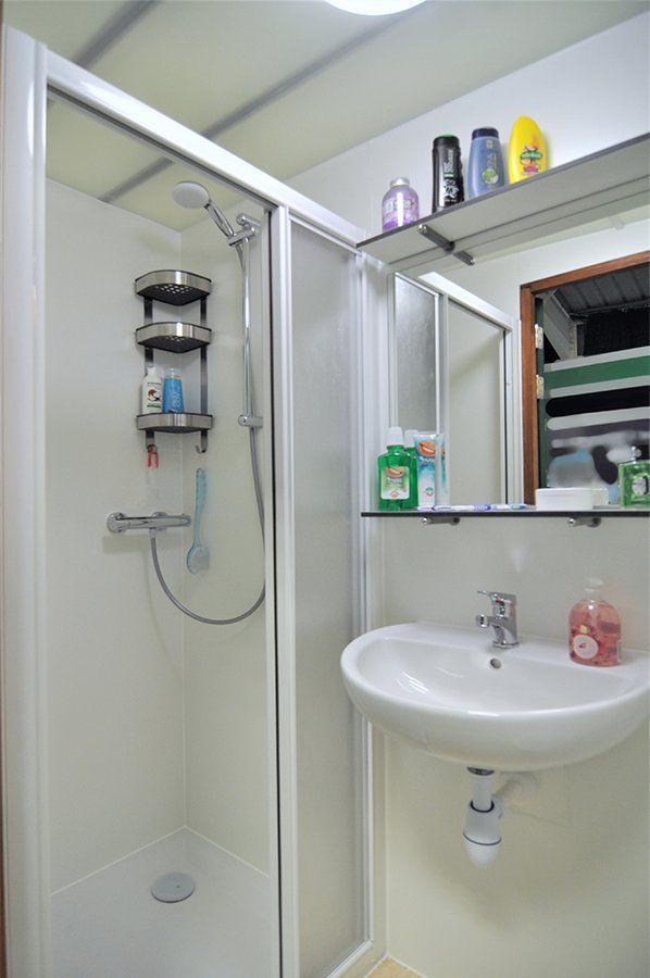 Kleine badkamer met douche, wastafel, spiegel en toiletartikelen op een plank en vensterbank.