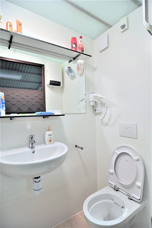 Kleine witte badkamer met wastafel, douche en open toiletbril onder een plank en spiegel.