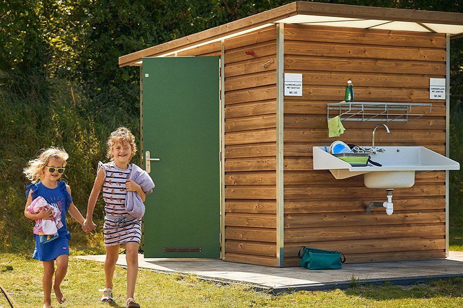 Twee kinderen staan naast een houten wasplaats in de buitenlucht, vlakbij een kleine hut.