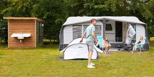 Een kampeertafereel met een gezin buiten een witte tent en een klein houten schuurtje op een grasveld.