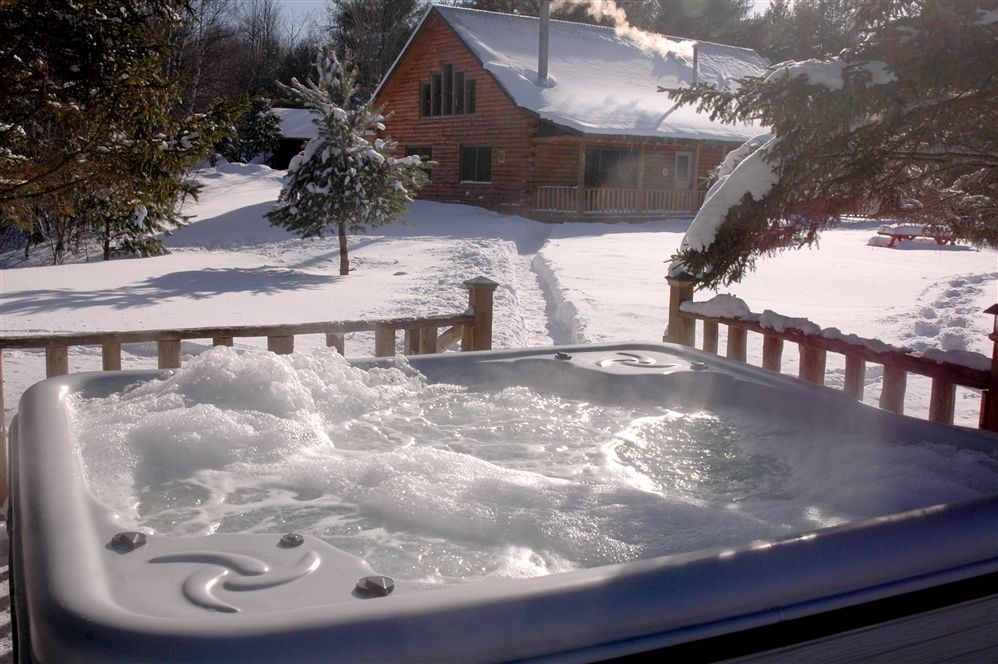 Snowy hot tub in a winter cabin yard with a log house and trees in the background