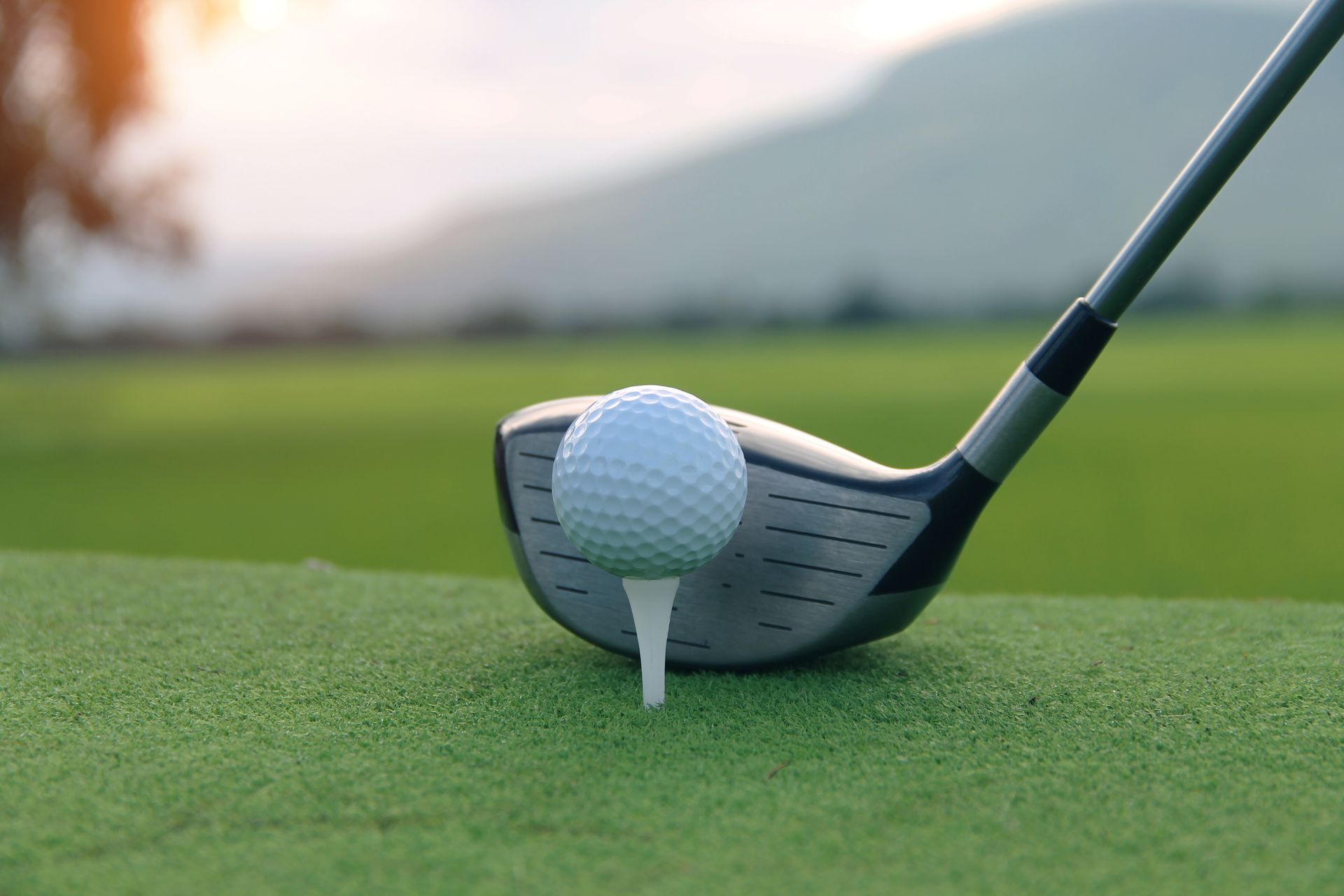 Golf ball on tee, driver approaching on green with blurred background of course and mountains.