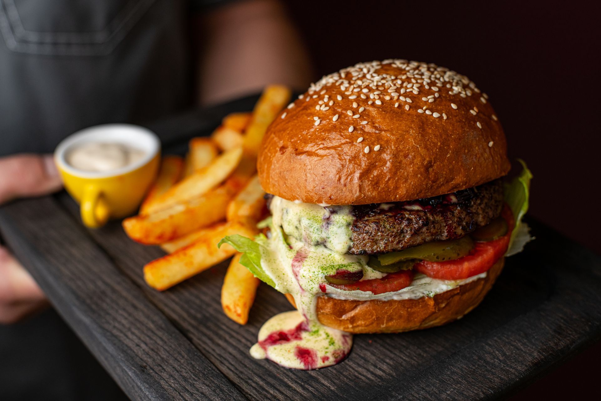 Burger with fries and dipping sauce on a wooden tray, held by a person.