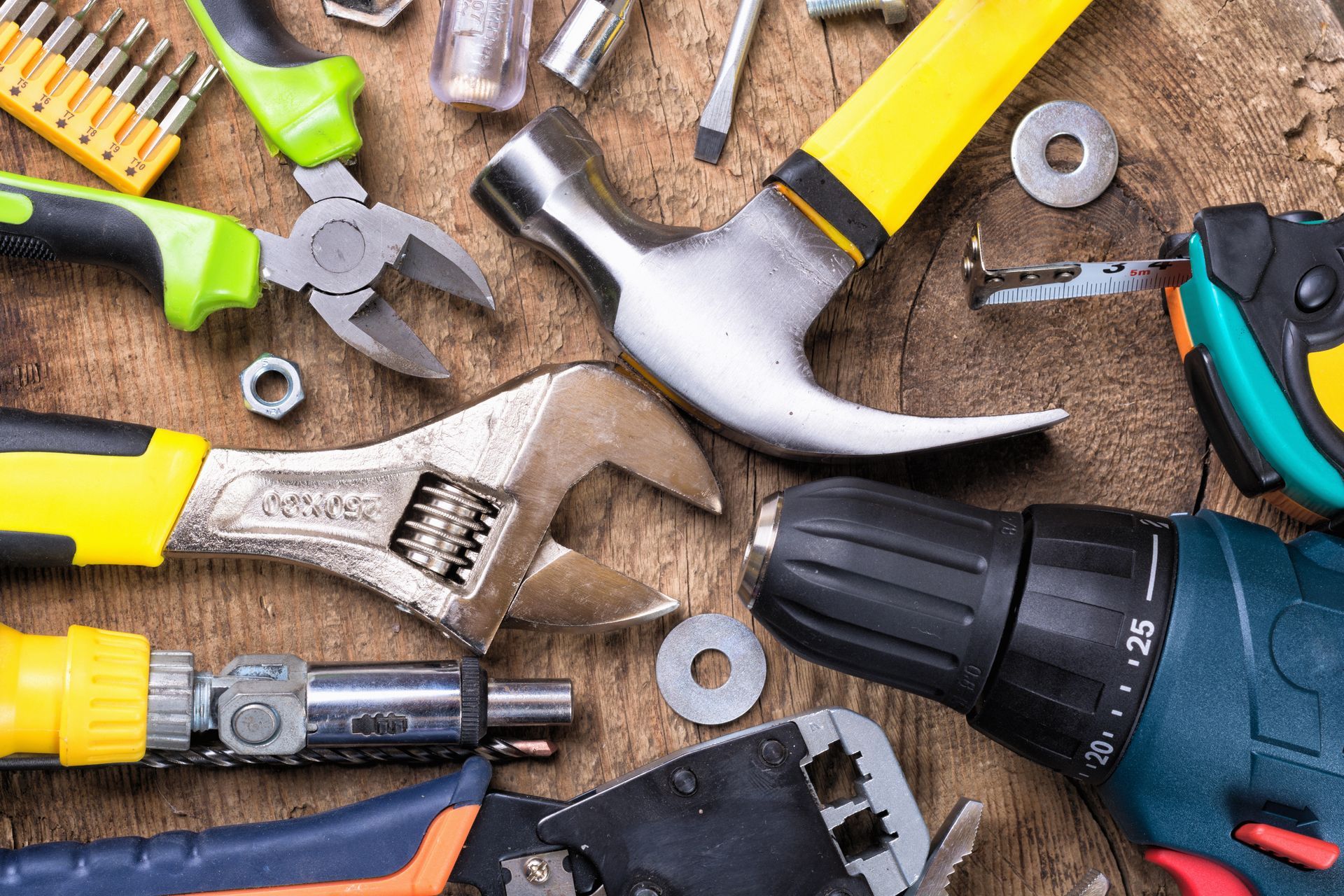 Assorted hand tools with yellow and green handles on a wooden workbench, including a hammer and drill