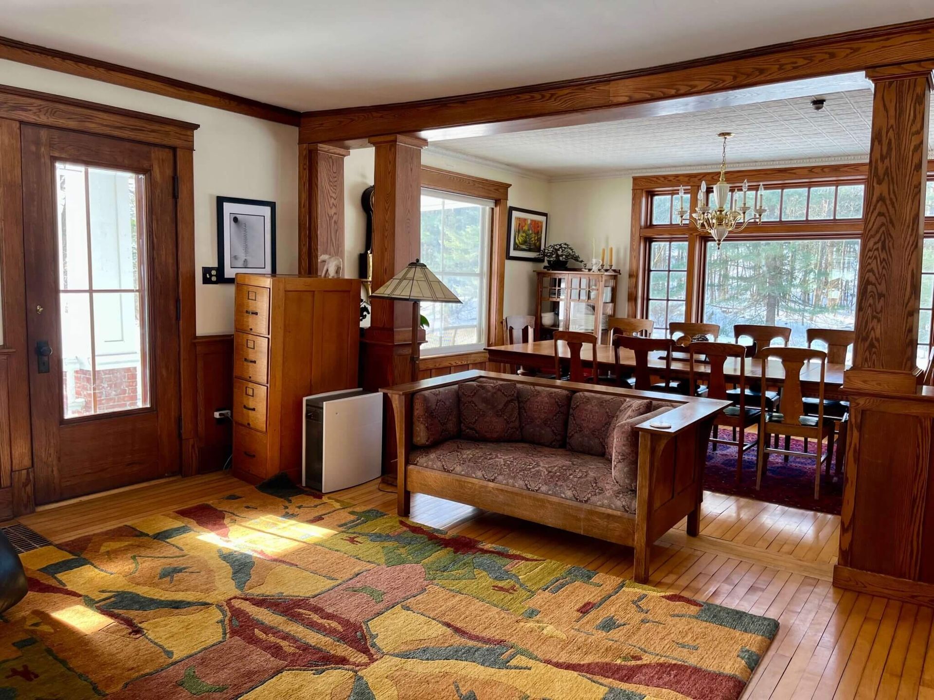 Living and dining room with hardwood floors, dark wood trim, rug, and furniture. Sunlight streams through the windows.