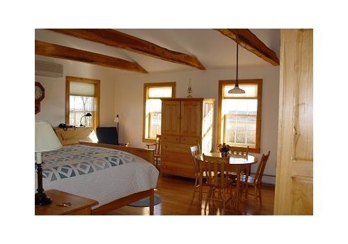 Cozy bedroom with wooden beams, bed, dresser, and small dining table by sunlit windows
