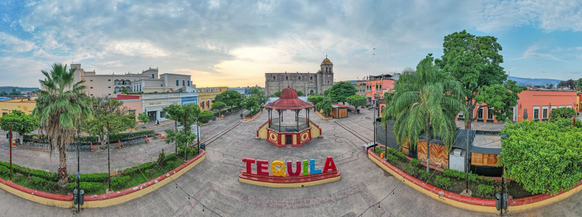 Amplia plaza con colorido letrero de “Tequila”, fuente, palmeras y edificios bajo un cielo nublado al atardecer.
