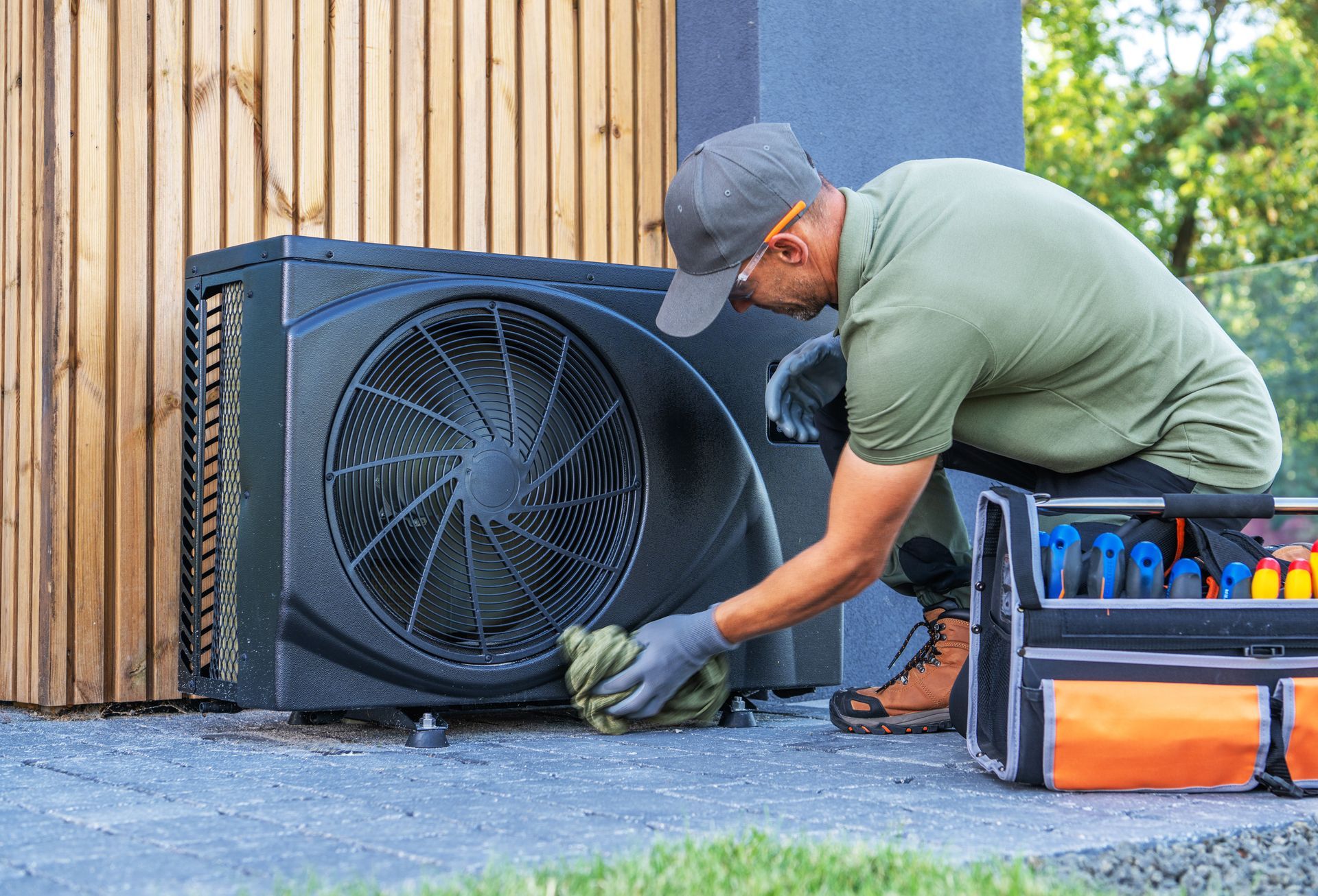 A technician wearing gloves cleans an outdoor heat pump unit next to a tool bag on a paved patio.