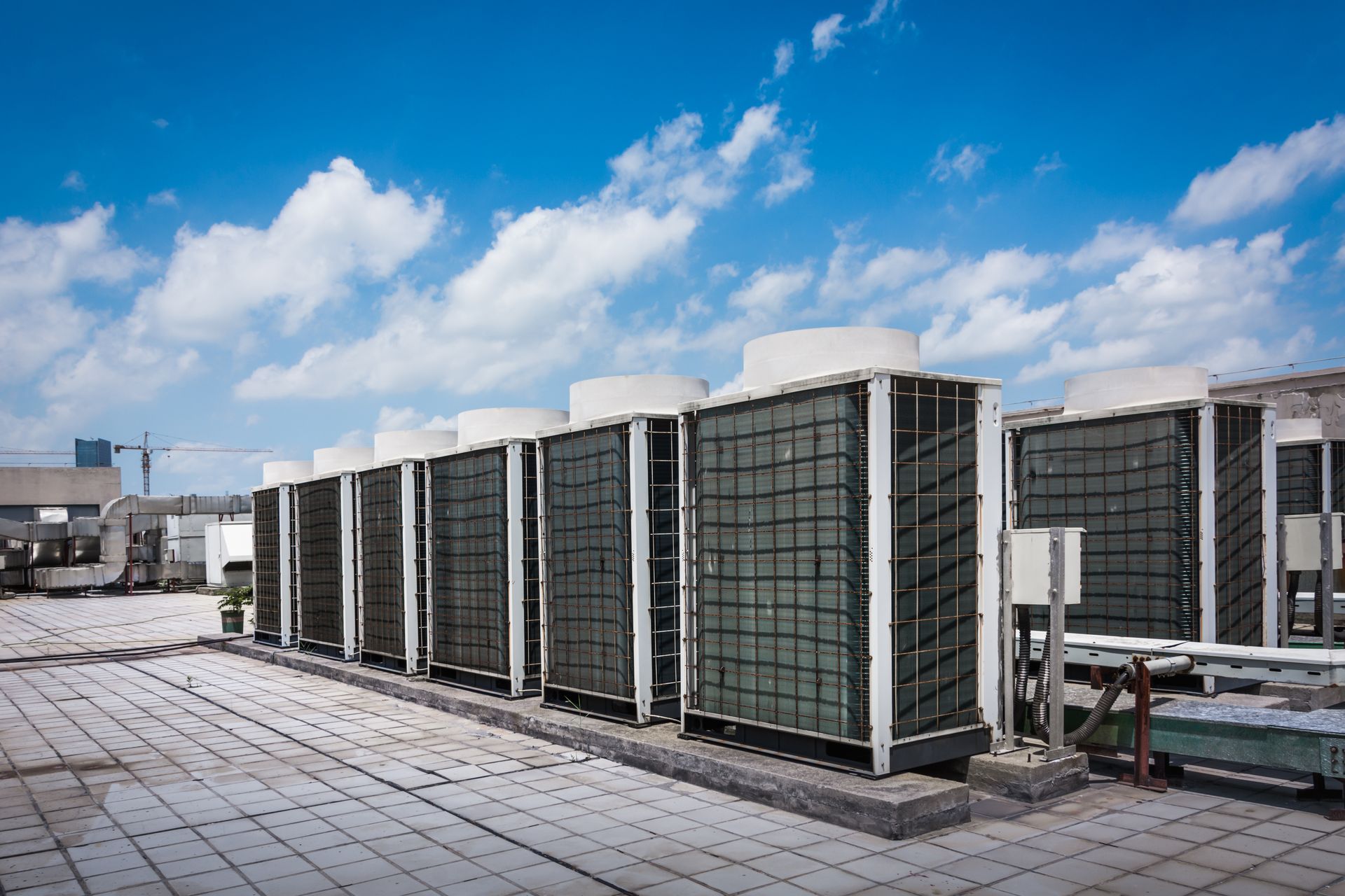 A row of industrial HVAC units on a rooftop under a blue sky with scattered clouds. A row of industrial HVAC units on a rooftop under a blue sky with scattered clouds.
