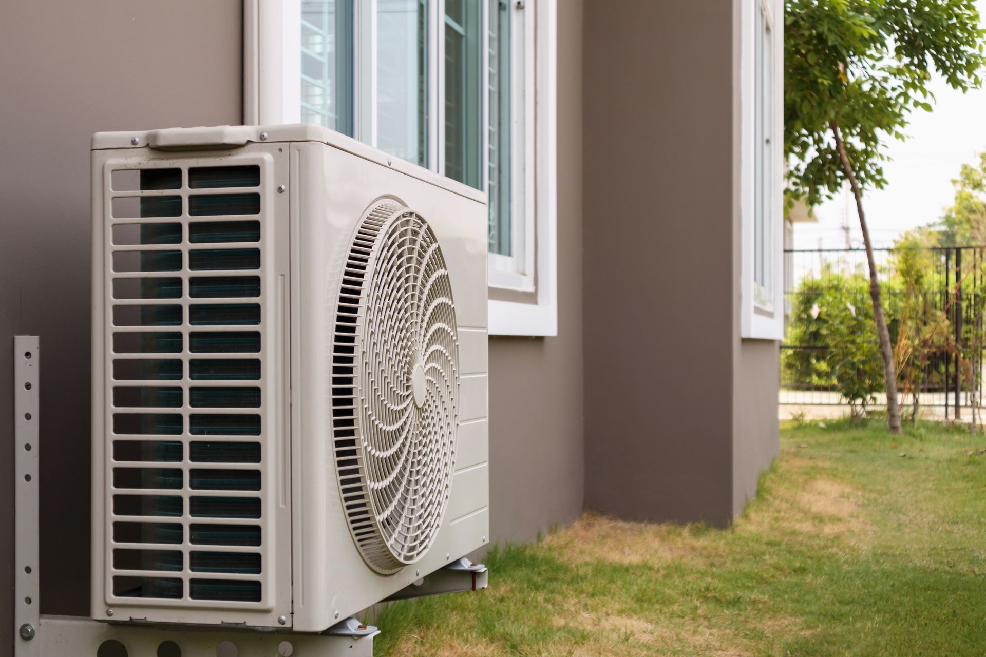 Air conditioning unit mounted on a brown house exterior, next to a window and green lawn.
