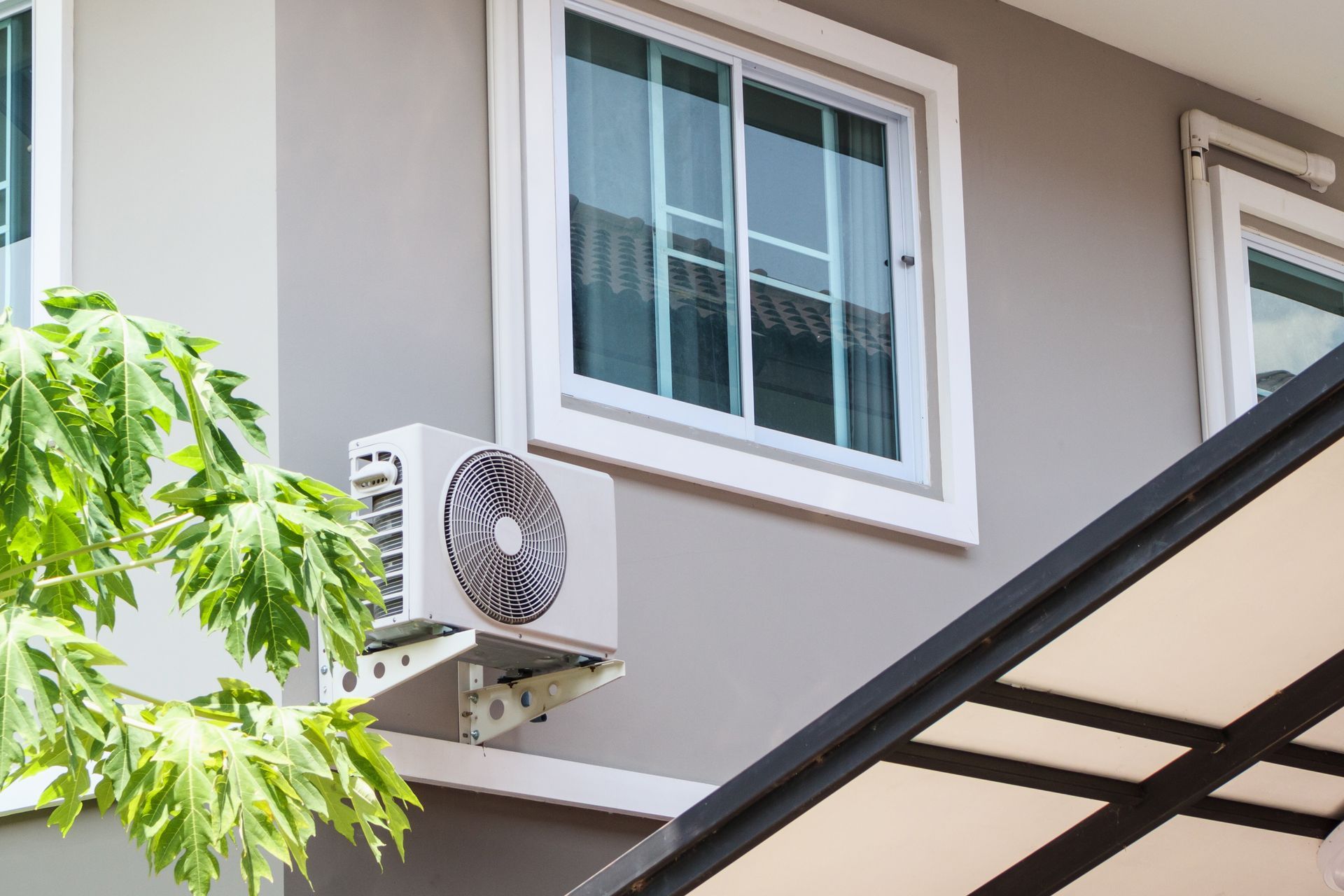 An outdoor air conditioning unit is mounted on a light gray exterior wall beneath a window, with a partial canopy nearby. An outdoor air conditioning unit is mounted on a light gray exterior wall beneath a window, with a partial canopy nearby.