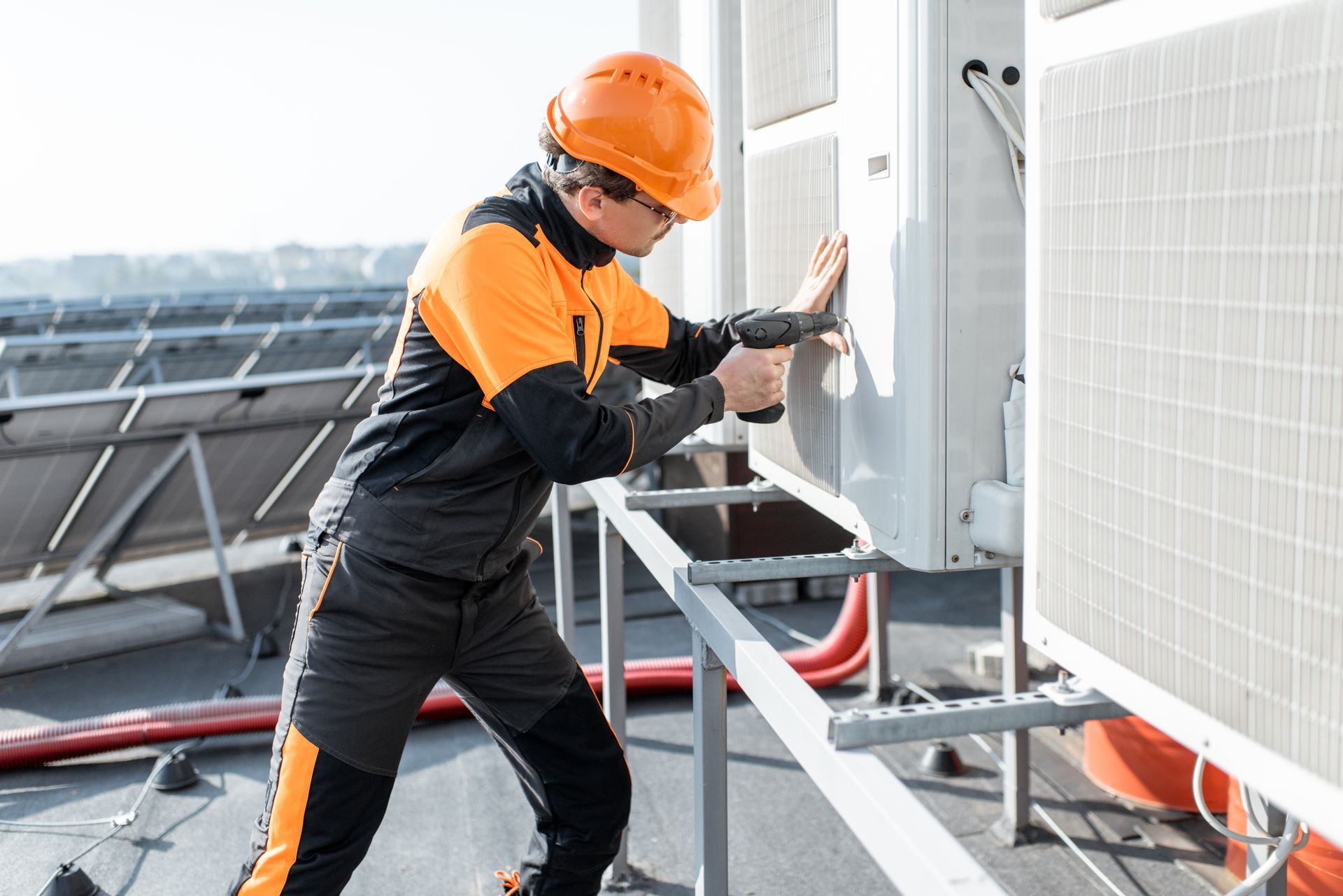 A worker in a high-visibility uniform and orange hard hat uses a drill on an outdoor HVAC unit on a rooftop.