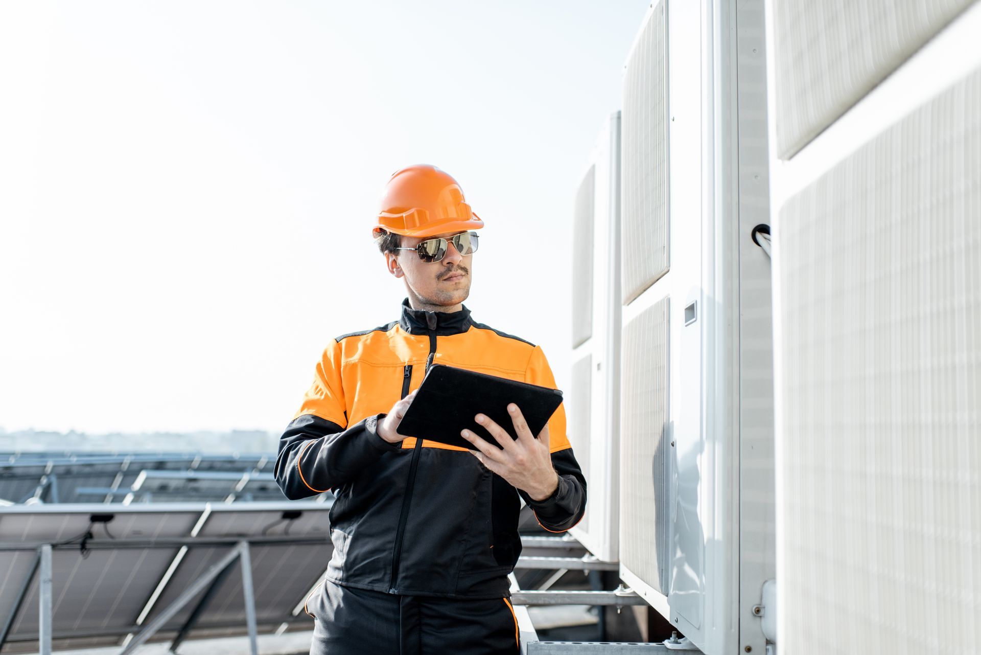 Technician in a hard hat and high-visibility jacket using a tablet to inspect HVAC units on a building roof. Technician in a hard hat and high-visibility jacket using a tablet to inspect HVAC units on a building roof.