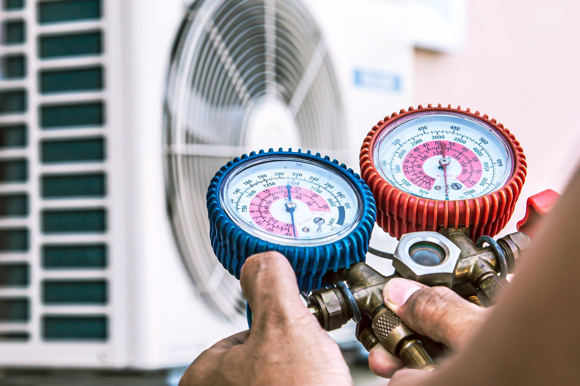 A technician holds blue and red HVAC manifold gauges in front of an outdoor air conditioning unit. A technician holds blue and red HVAC manifold gauges in front of an outdoor air conditioning unit.
