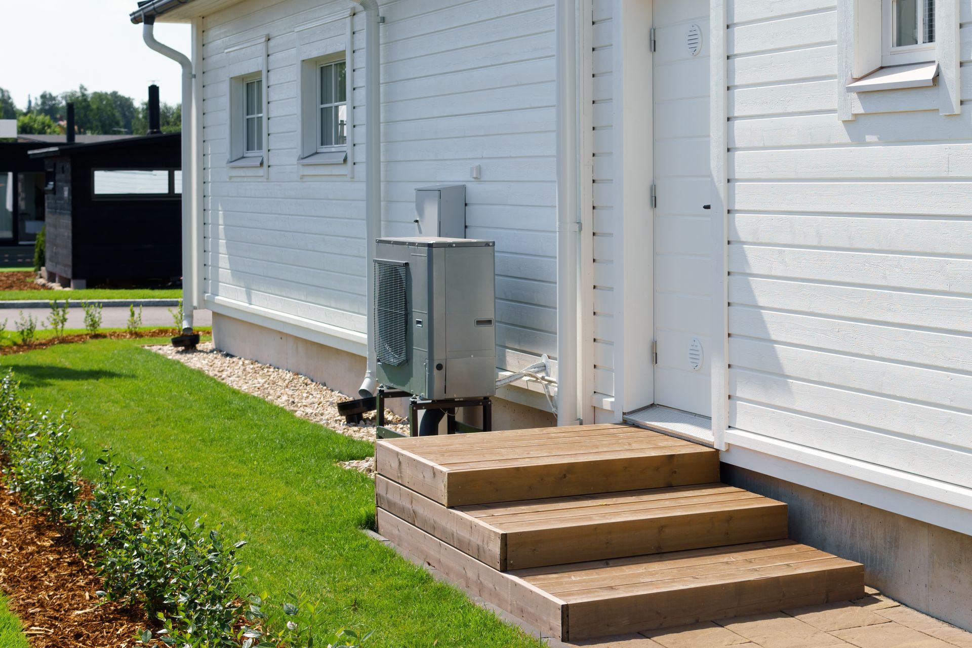 A gray heat pump unit stands beside the wooden steps leading to a white, horizontal-sided house with a manicured lawn.