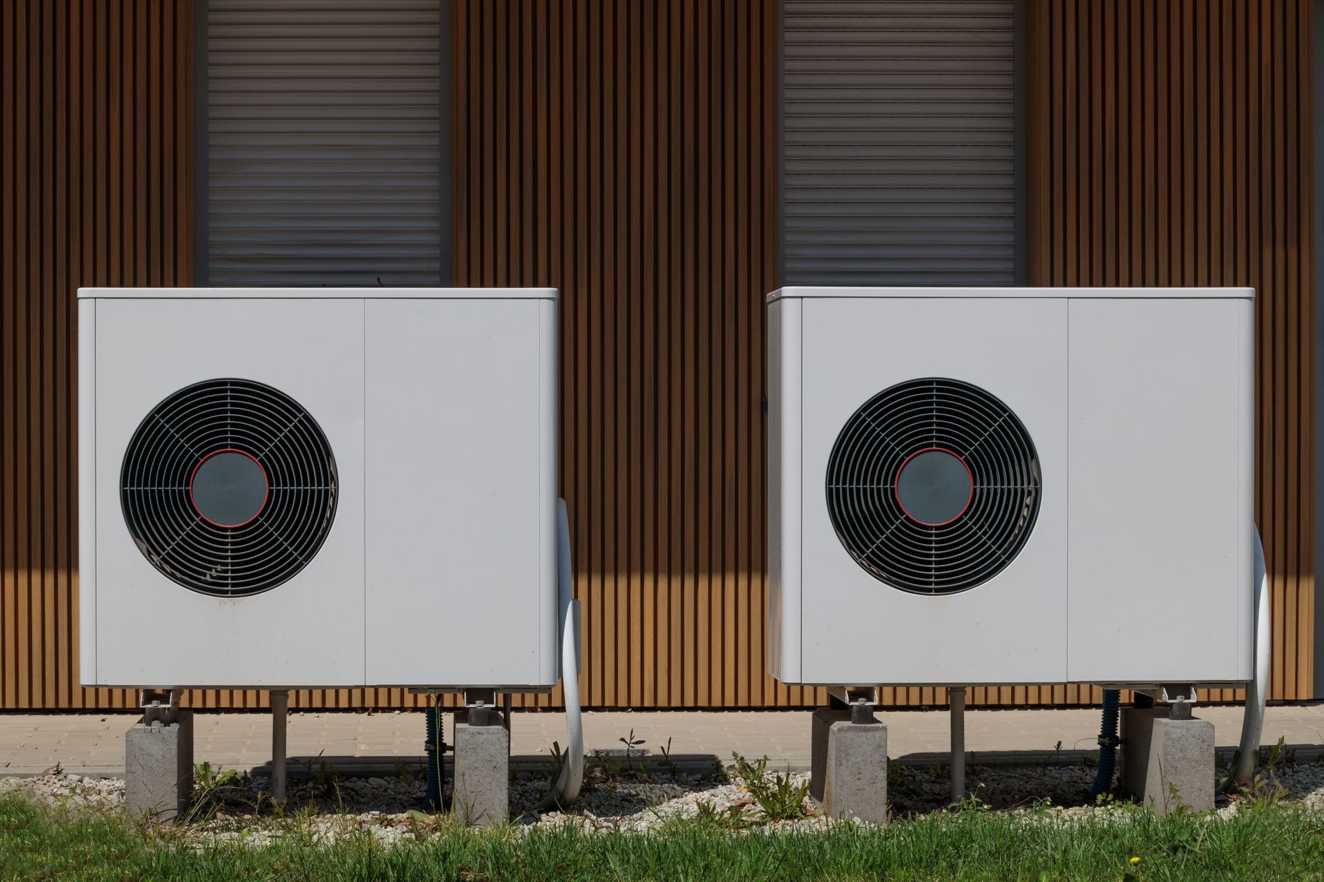 Two white heat pump units sit on concrete blocks against a brown, vertically slatted building wall. Two white heat pump units sit on concrete blocks against a brown, vertically slatted building wall.