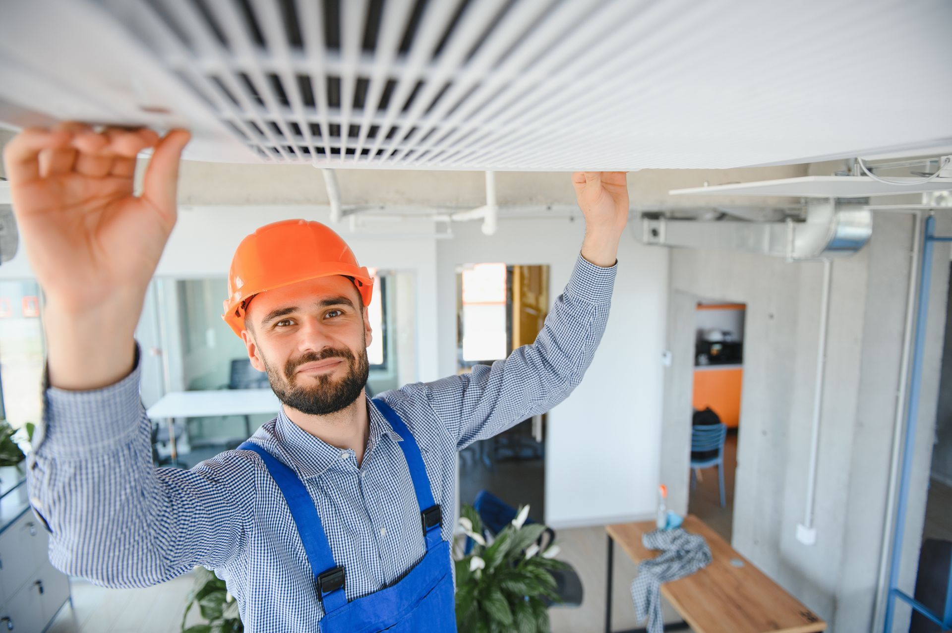 A person in an orange hard hat and blue coveralls installing a ventilation grate on a ceiling in a modern office space.