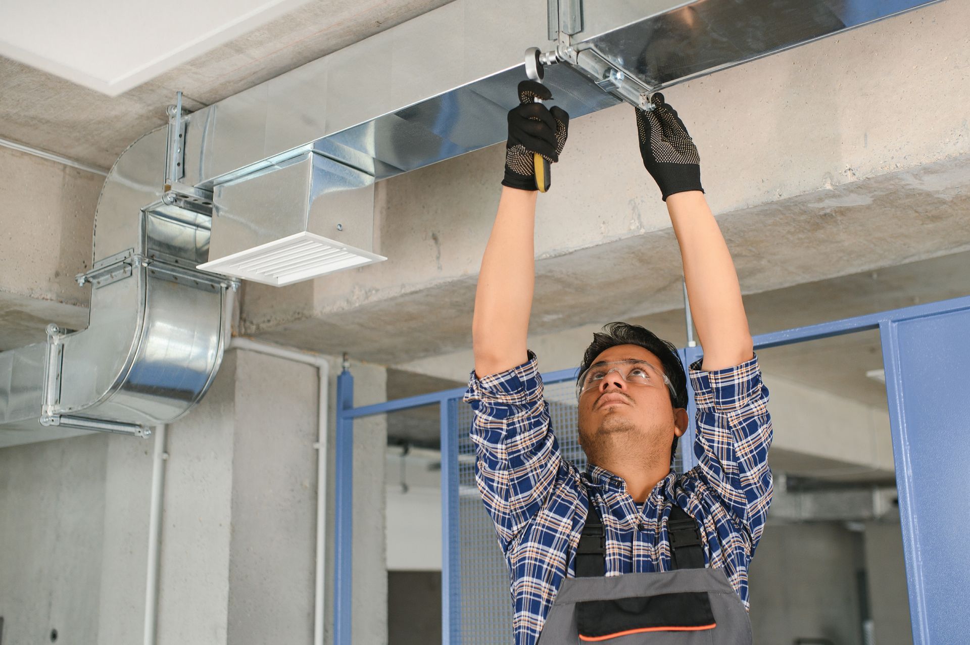 Man installing HVAC ductwork in a building. He wears gloves, overalls, and looks up.