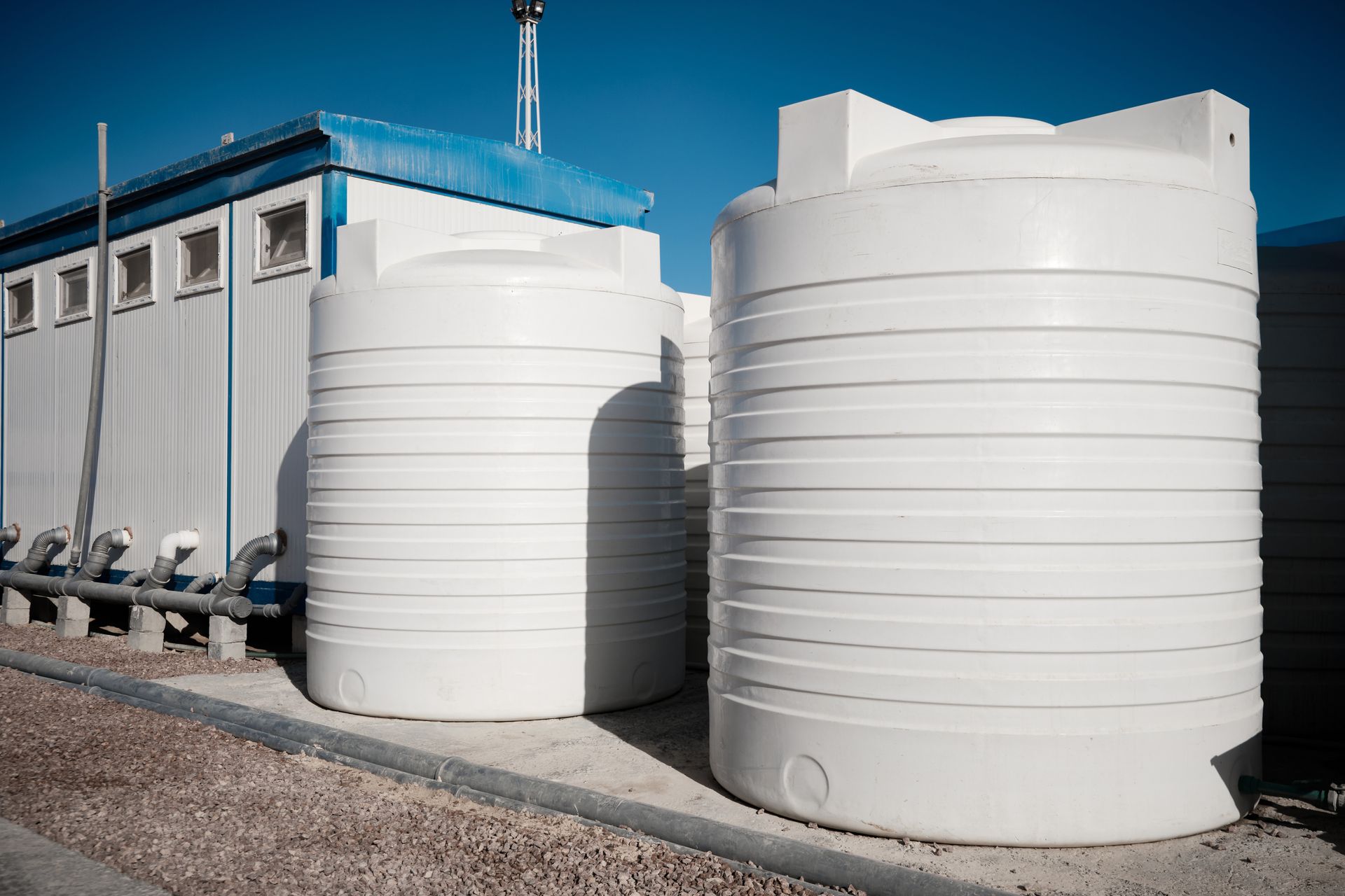 Large white water storage tanks beside a building on a gravel lot under a clear blue sky Large white water storage tanks beside a building on a gravel lot under a clear blue sky