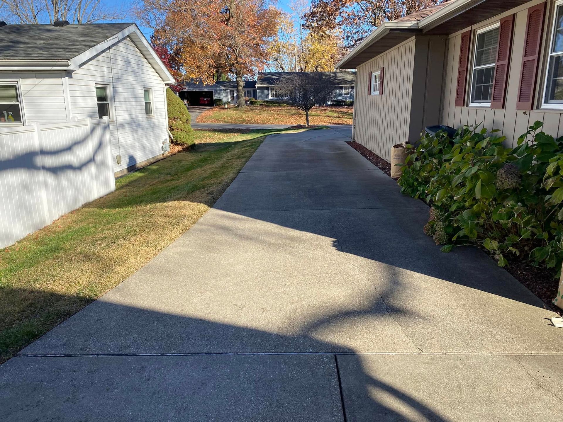 A concrete driveway between two houses with a garage and autumn trees in the background.
