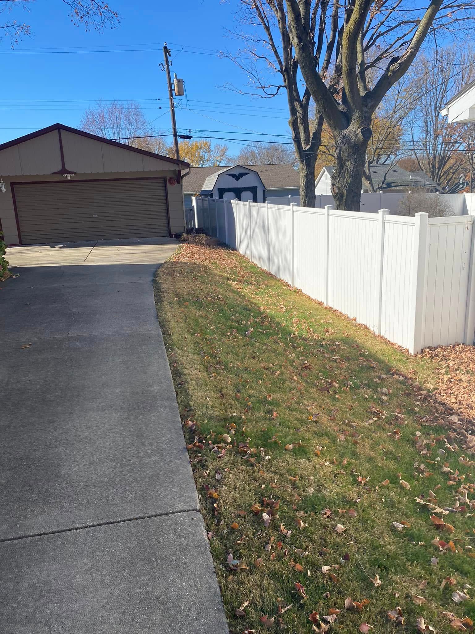 Driveway beside white fence and grassy strip leading to a garage under a clear blue sky.
