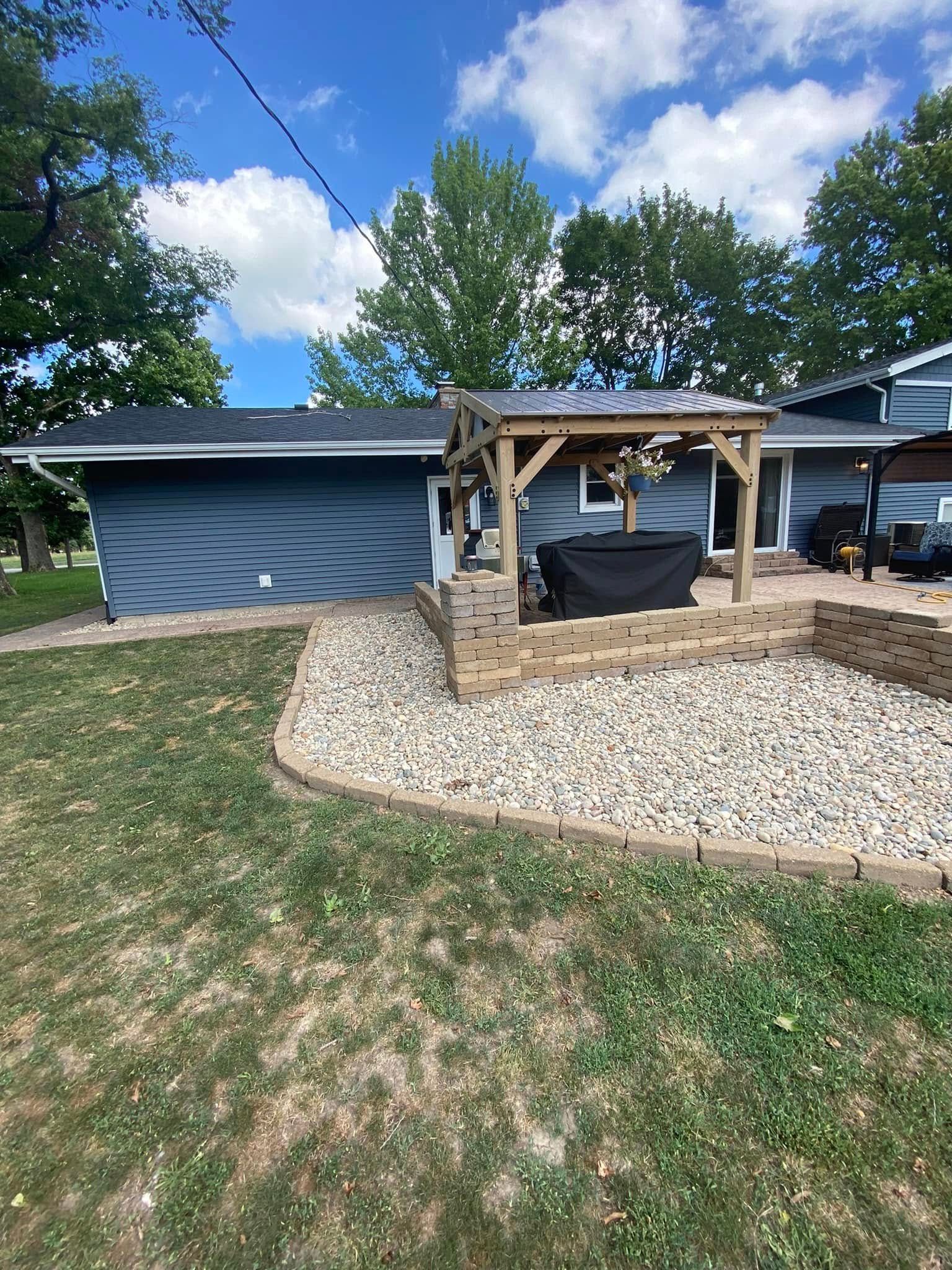 Backyard patio with a wooden pergola swing, gravel border, curved blue shed, and grassy lawn under a cloudy sky