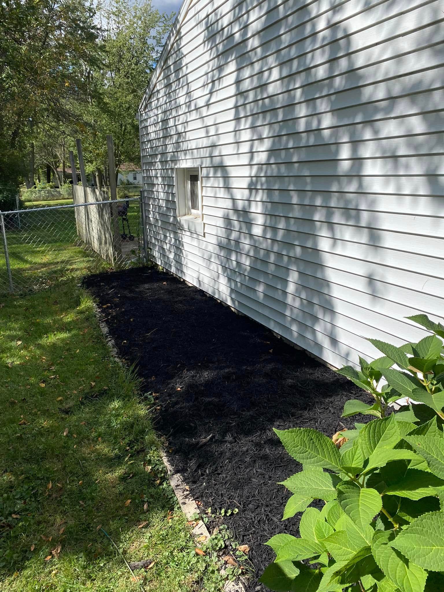 White house siding beside a narrow black mulch bed and green lawn, with trees and a fence in the background