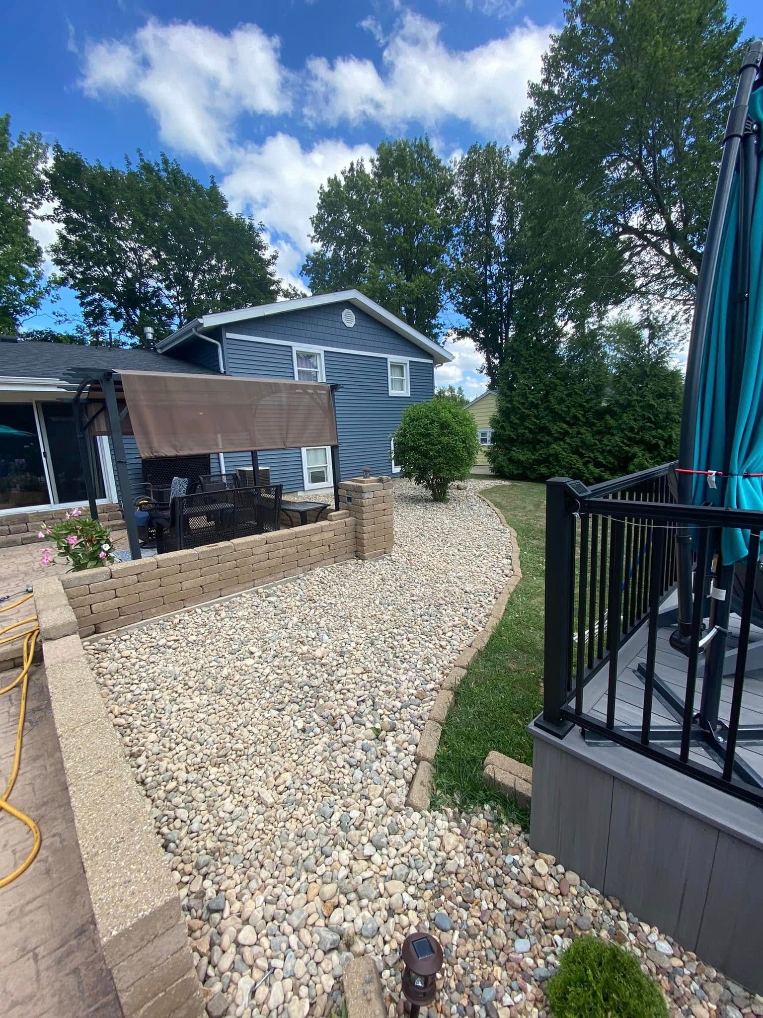Gravel pathway beside a house with a stone retaining wall, black railing, and blue sky with trees.