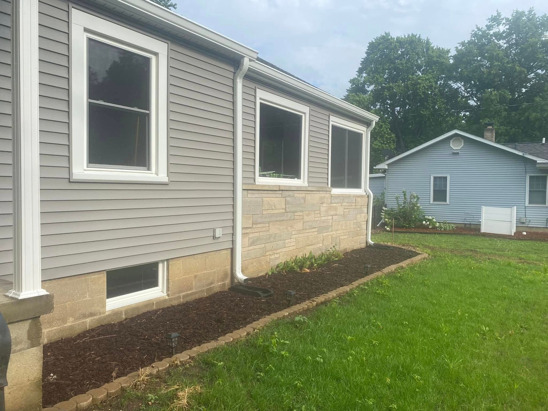 Side of a house with beige siding and stone veneer, windows, and a green lawn under a cloudy sky.
