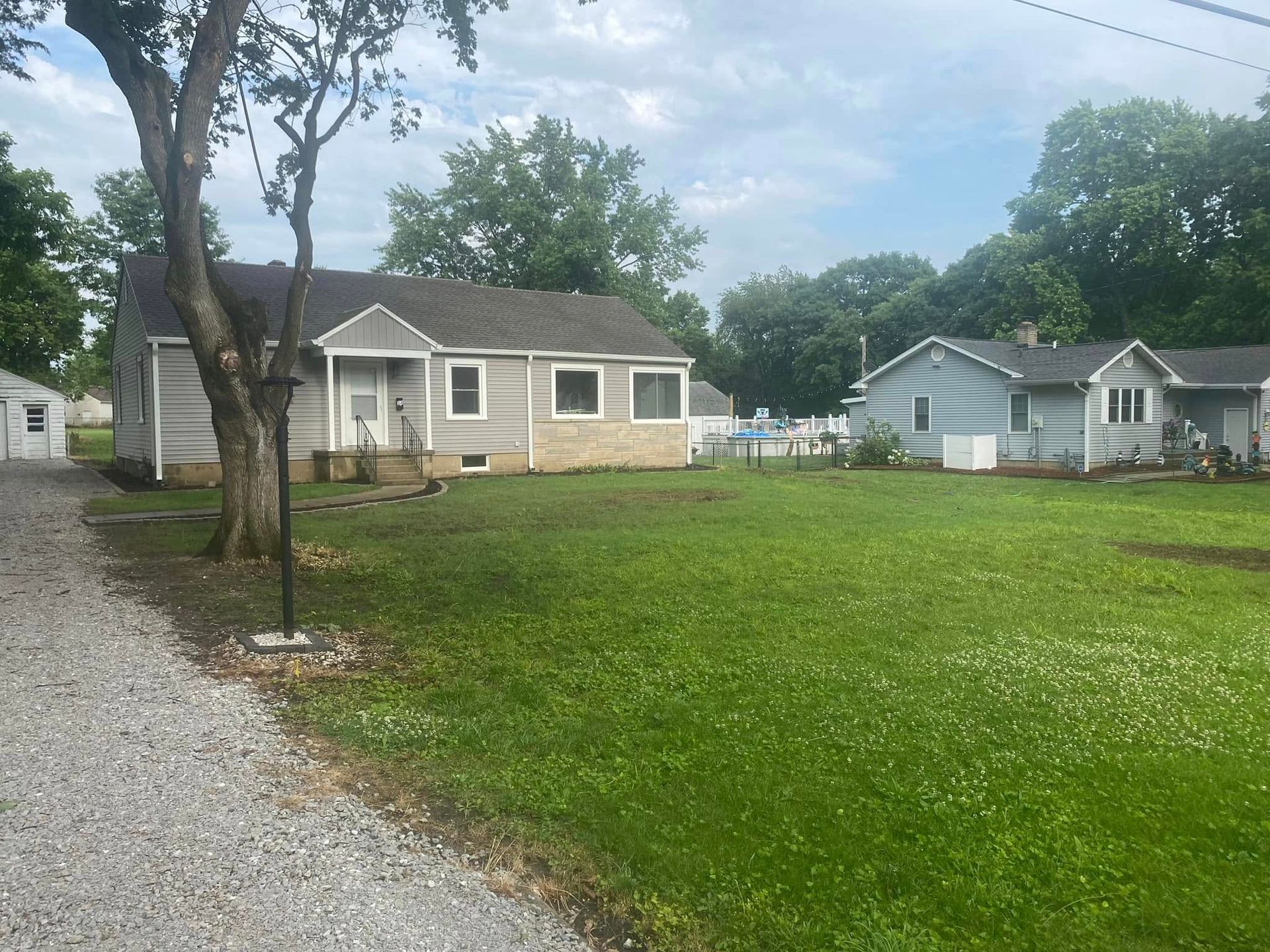 Small gray house with stone base beside a gravel driveway and grassy yard under cloudy skies