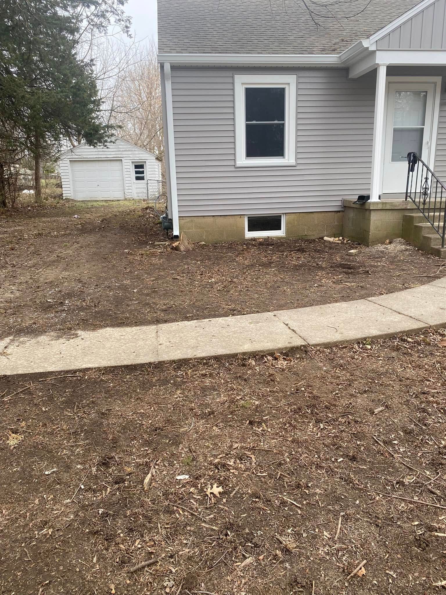 Gray house exterior with leaf-covered yard, curved concrete walkway, and a small basement window