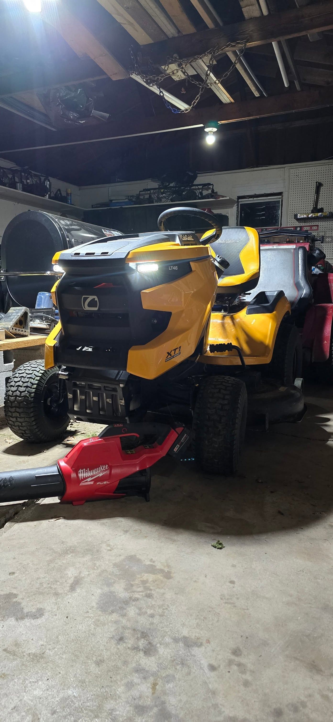 Yellow riding lawn mower in a dim garage, with a red snowblower attachment in front.