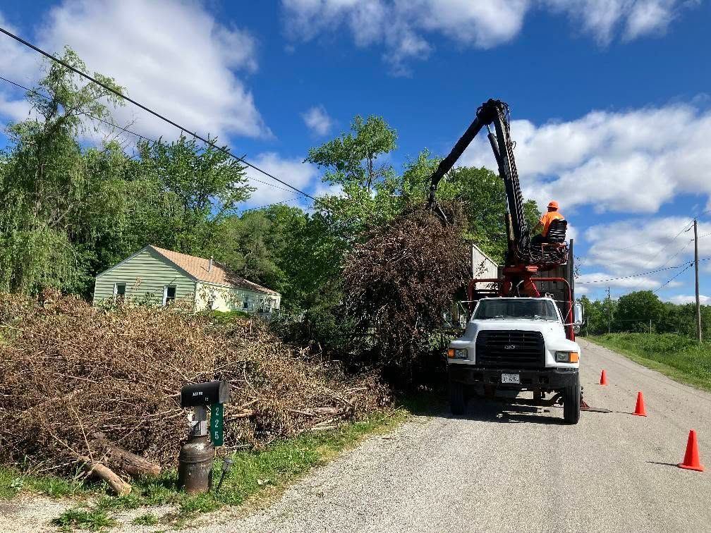Utility crew clearing storm debris beside a road, with a crane truck and orange cones under a blue sky.