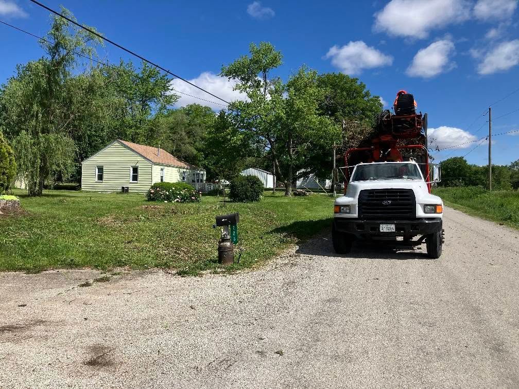 White truck loaded with equipment parked on a gravel road near a small house and trees under a blue sky
