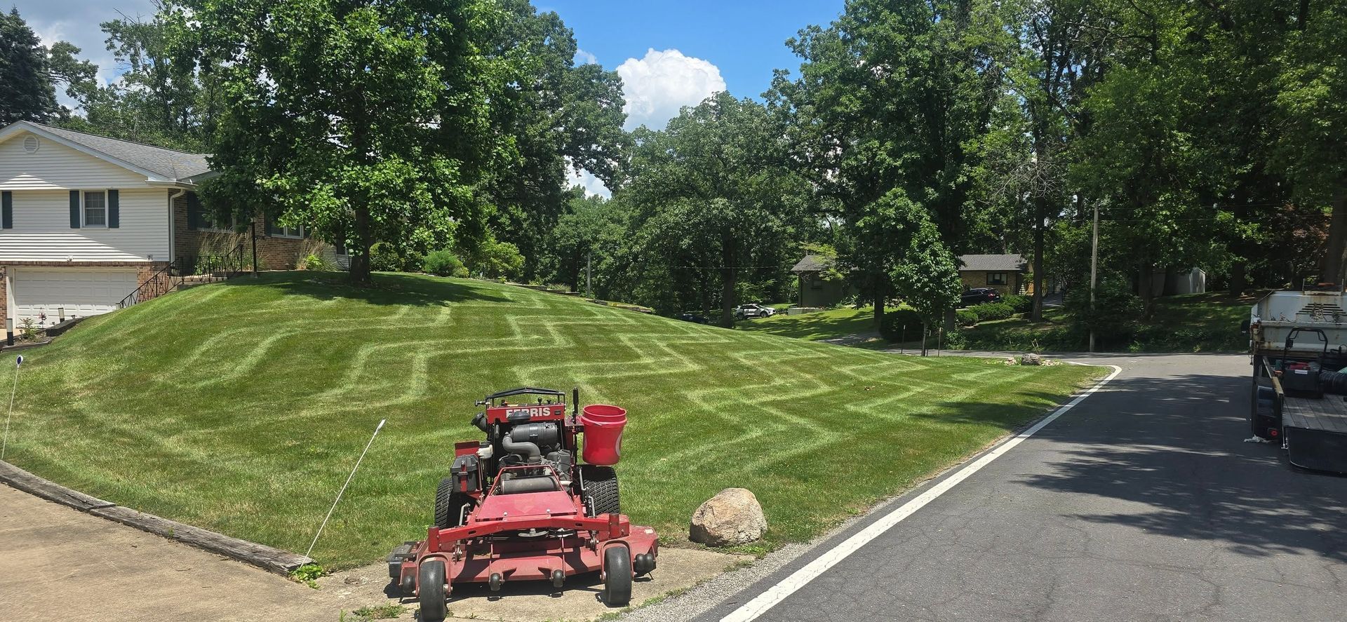 Red riding lawn mower on a mowed grassy hill beside a driveway and parked vehicle