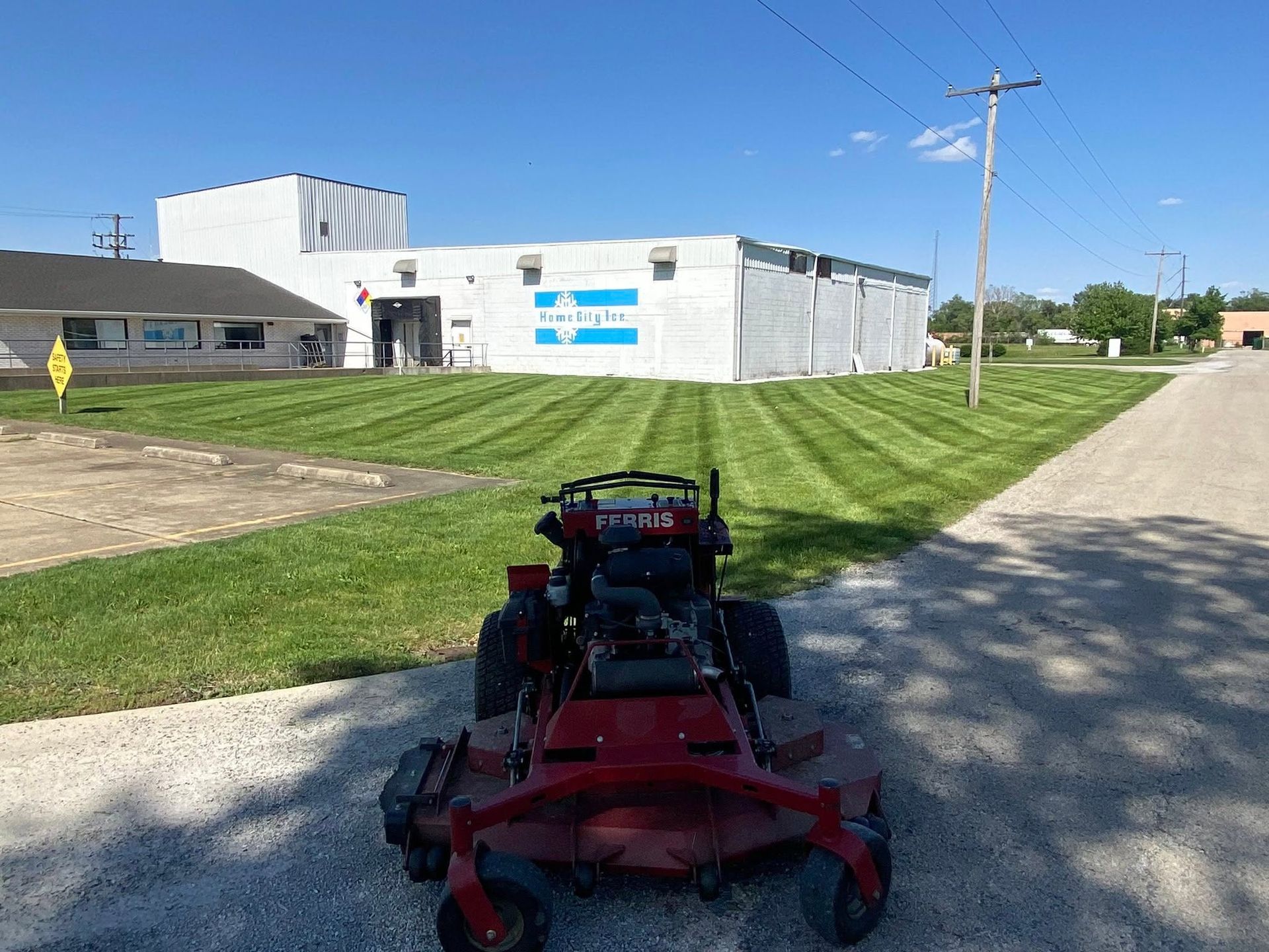 Red lawn mower on grassy roadside beside a white industrial building under a clear blue sky