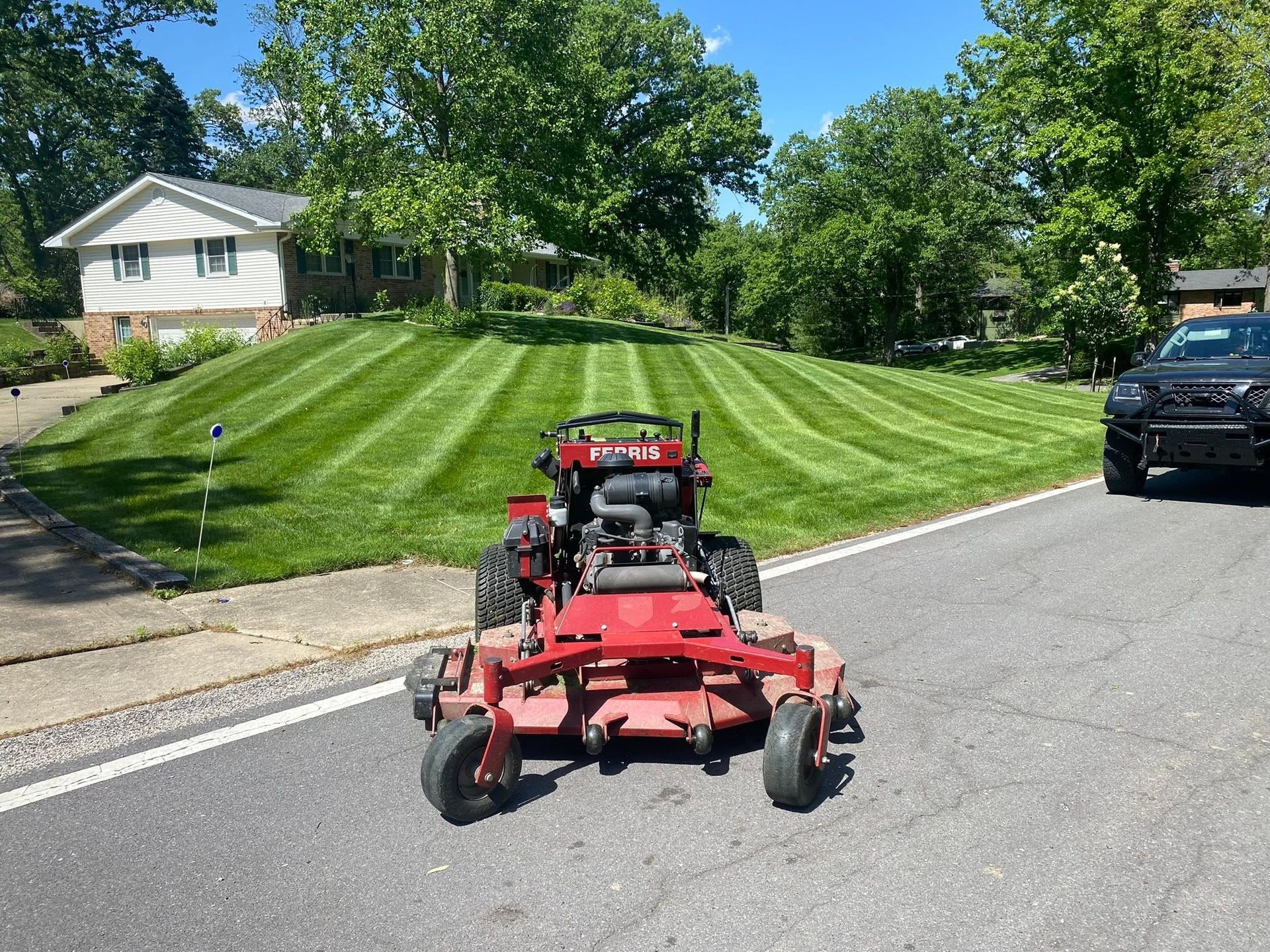 Red riding lawn mower on a striped grassy slope beside a street, with houses and parked cars nearby