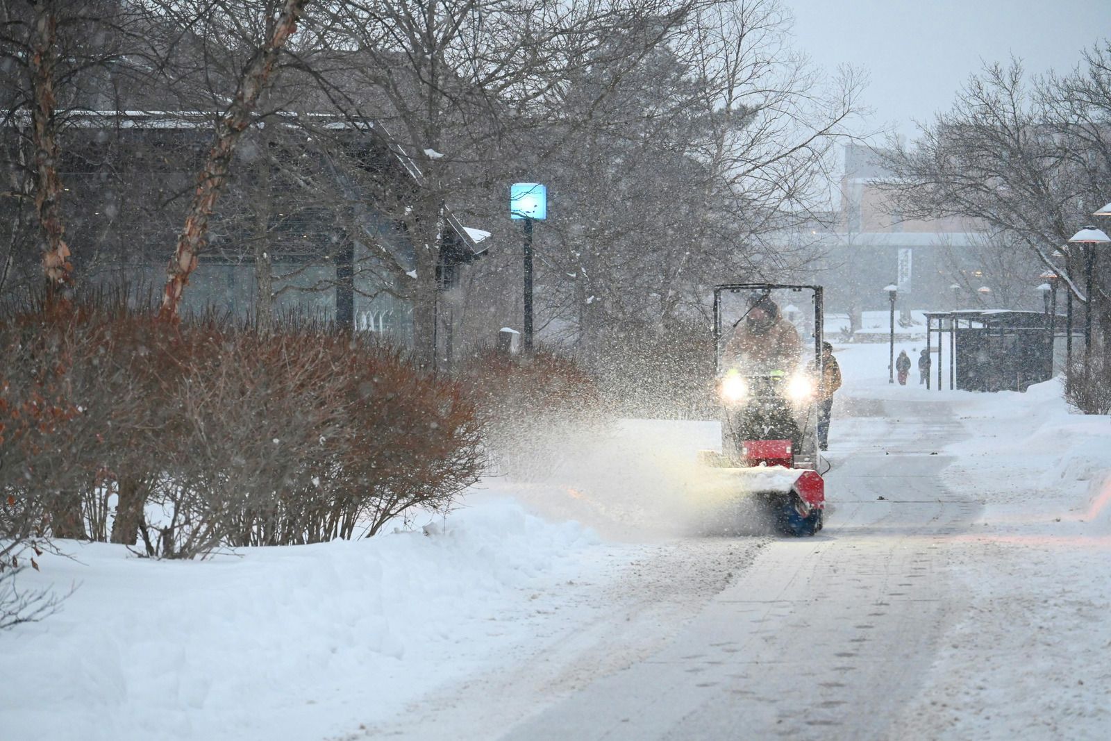 Snowplow driving down a snowy residential street during snowfall, headlights on.