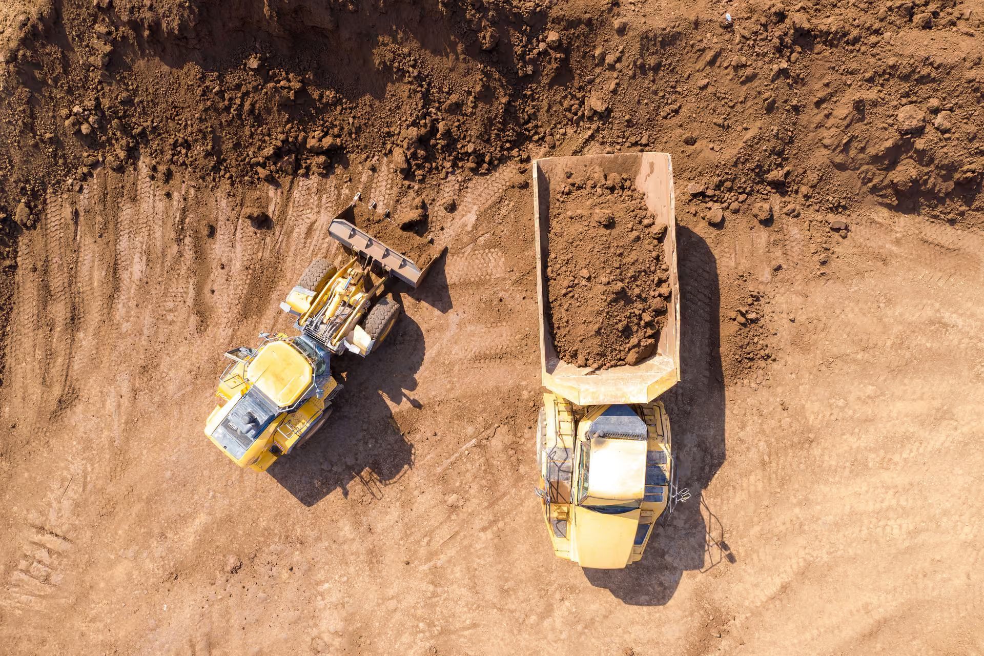 Aerial view of two yellow construction dump trucks on a dirt site, one loading soil and one full of earth